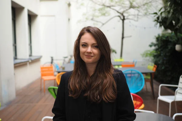 Sophie Riche, cheveux bruns mi-longs, vêtue d'un haut noir, debout dans une cour extérieure avec terrasse en bois et chaises colorées, souriant légèrement.