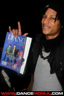 Larry des Twins, souriant avec un bonnet noir, tenant un exemplaire du magazine 'Dance Mogul Magazine' lors du festival international de street dance Juste Debout, avec un filigrane URL en bas de l'image.