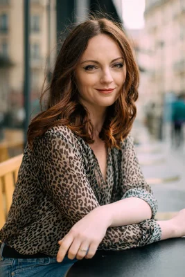 Sophie Riche, cheveux bruns ondulés, portant un chemisier léopard transparent, appuyée sur une table sombre dans un décor urbain flou.