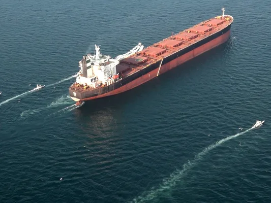 Un pétrolier turc et un autre japonais traversant le détroit d'Ormuz, avec des navires militaires en arrière-plan, illustrant les tensions dans la région.