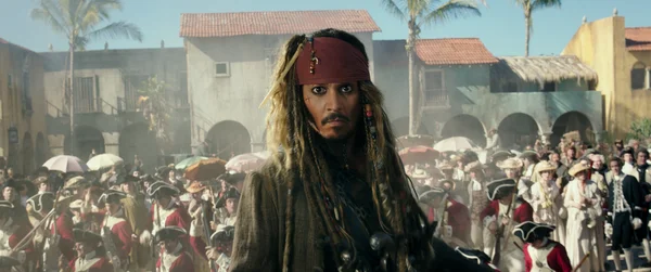 Johnny Depp en Jack Sparrow, avec des dreadlocks longues et sombres, un bandana rouge et du khôl, dans une place de ville coloniale dans Pirates des Caraïbes : La Mort ne attend pas (2017).