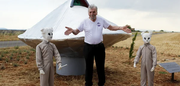 Un homme les bras écartés devant une réplique argentée d'OVNI en forme de soucoupe, entouré de deux figurants en combinaison grise et masque d'alien blanc, lors de la célébration des 60 ans de la rencontre du troisième type à Valensole.