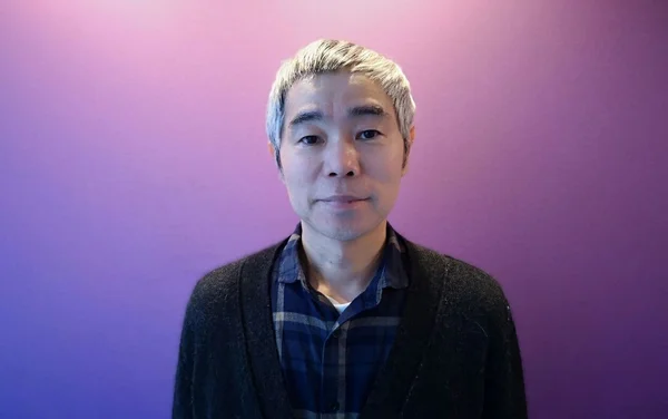 Portrait en buste de Taiyô Matsumoto, mangaka japonais, avec des cheveux courts argentés, portant un gilet sombre sur une chemise à carreaux bleue, sur un fond dégradé bleu-violet.