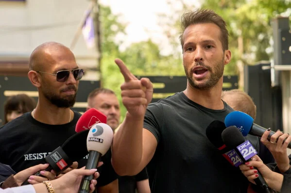 Andrew Tate, influenceur aux cheveux bruns courts et à la barbe, s'adressant à des journalistes en levant la main droite, accompagné d'un homme portant des lunettes de soleil.