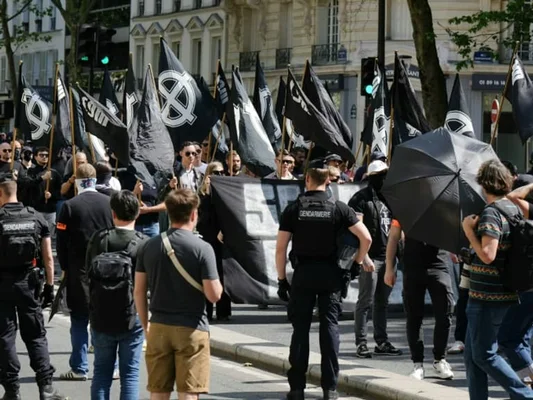 Manifestants marchant dans une rue parisienne avec des drapeaux noirs portant un symbole blanc circulaire, sous la surveillance de gendarmes français lors d'une marche néonazie du Comité du 9 Mai.