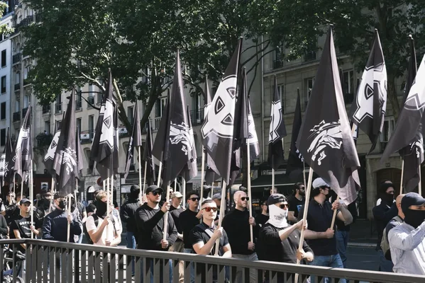 Groupe de néonazis défilant dans une rue parisienne, portant des drapeaux noirs avec des symboles blancs dont une croix celtique et une tête de loup, certains individus le visage dissimulé.