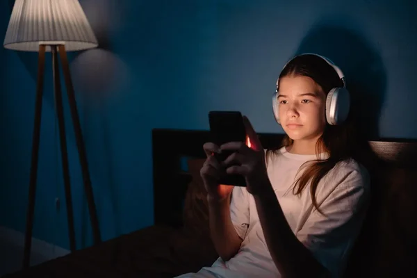 Une adolescente portant des écouteurs blancs regarde son smartphone dans une pièce sombre, illustrant la consommation d'information en ligne par les jeunes