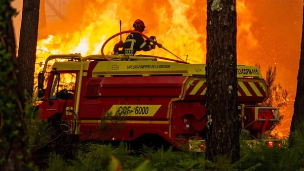 Un pompier sur un camion de pompiers rouge utilise un canon à eau pour combattre un incendie de forêt massif en Gironde, avec des flammes intenses en arrière-plan