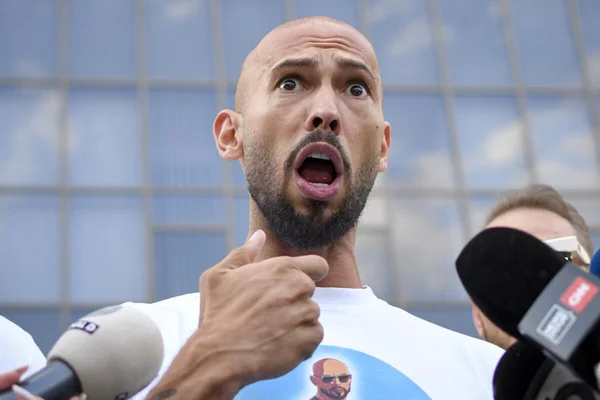 Andrew Tate, influenceur des réseaux sociaux, barbu et rasé de près, s'adressant à un groupe de journalistes avec des microphones devant un bâtiment moderne en verre
