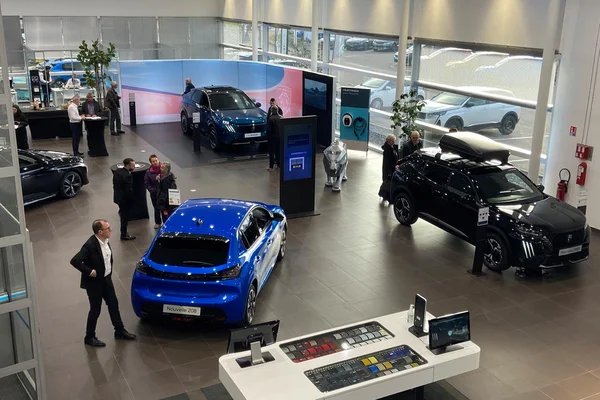 Intérieur d'un concessionnaire automobile moderne avec plusieurs véhicules dont une Peugeot 208 bleue et un SUV noir, des clients et du personnel interagissant dans l'espace de vente
