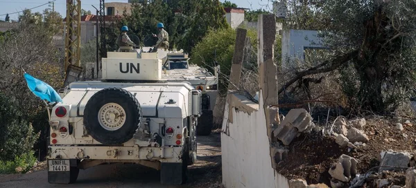 Un véhicule blindé de transport de troupes des Nations Unies avec deux soldats en casques bleus circulant sur une route étroite du sud du Liban, à côté d'un mur en béton endommagé