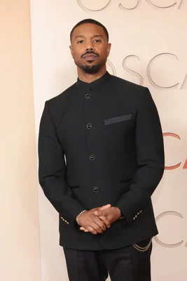 Michael B. Jordan portant une veste à col mandarin noire lors de la cérémonie des Oscars 2026, avec les mains jointes devant un arrière-plan clair.