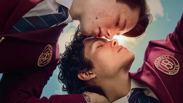 Deux jeunes hommes en uniforme scolaire bordeaux se tenant de près dans une pose intime sous un ciel éclatant, extrait de la série Young Royals.