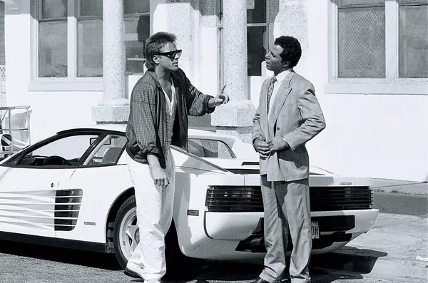 Deux personnages de la série Miami Vice, l'un en chemise à carreaux et lunettes de soleil, l'autre en costume, discutant à côté d'une blanche Ferrari Testarossa emblématique.