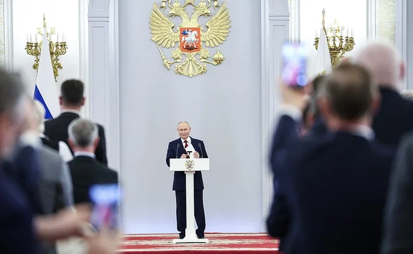 Le président russe Vladimir Poutine en costume sombre s'adressant à des députés de la Douma depuis un podium, avec l'emblème à double tête d'aigle doré en arrière-plan.