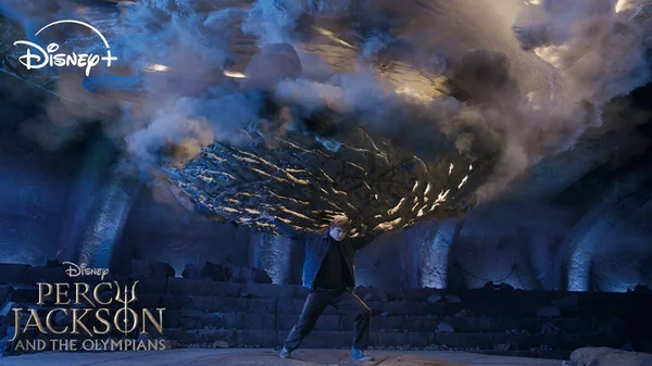 Walker Scobell en Percy Jackson portant un sweat-shirt bleu, soulevant une énorme dalle de pierre dans un environnement souterrain enfumé