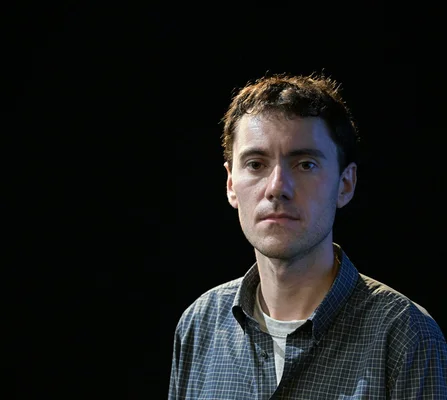 Portrait de Robin Josserand, auteur du roman 'Après Mireille', en chemise à carreaux bleus et blancs sur fond noir