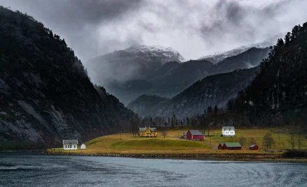 Vue panoramique d'un petit village suédois aux maisons colorées situé sur une berge herbeuse le long d'un fjord, entouré de montagnes escarpées aux sommets enneigés sous un ciel nuageux.