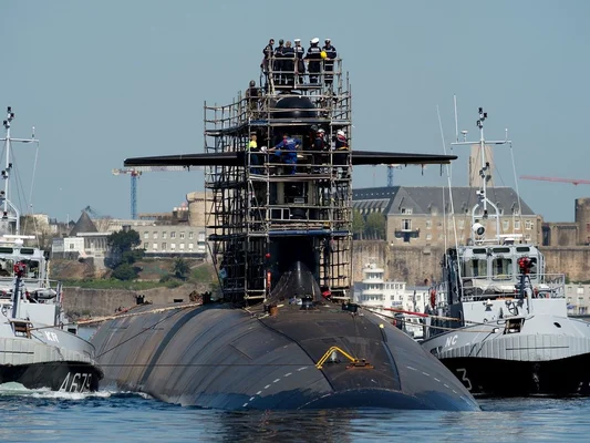 Sous-marin noir de la base de l'Île Longue manœuvré par deux remorqueurs dans la rade de Brest, avec des échafaudages autour du kiosque où se tiennent plusieurs personnes