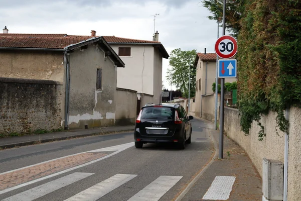 Photo montrant une rue étroite dans un village de la Métropole de Lyon avec une voiture noire approchant d'un panneau de limitation de vitesse à 30 km/h et des bâtiments en pierre ancienne.