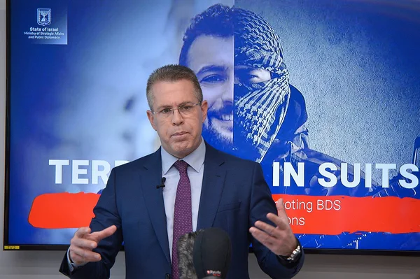 Un homme en costume sombre et cravate s'adressant à une tribune, devant un grand écran affichant le logo du ministère israélien des Affaires stratégiques et de la diplomatie publique, avec des textes comme 'TERROR IN SUITS' et 'Promoting BDS'.