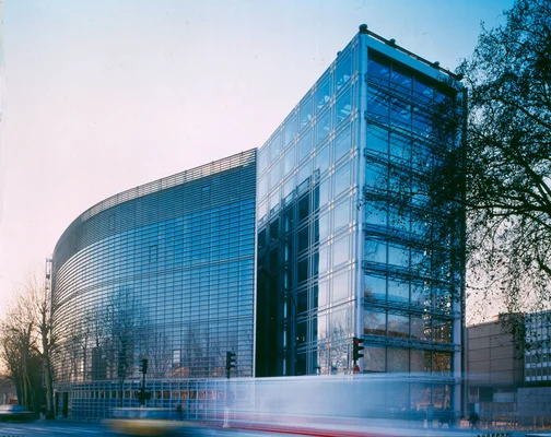 Vue en pose longue de l'Institut du Monde Arabe, bâtiment moderne aux formes angulaires et vitrées au crépuscule, avec traînées de lumière de véhicules en mouvement.