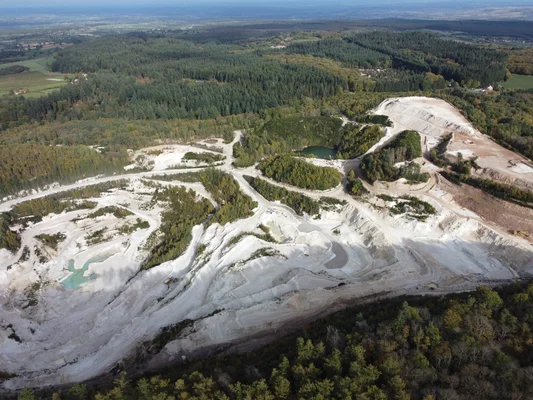 Vue aérienne d'une grande carrière à ciel ouvert aux terres claires et pentes en terrasses, avec un petit lac turquoise, entourée de forêt dense et de collines.