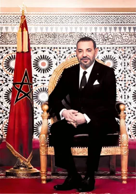Sa Majesté le Roi Mohammed VI en costume noir et cravate, assis sur une chaise dorée ornée à côté du drapeau marocain rouge, devant un fond de motifs géométriques et calligraphie arabe.