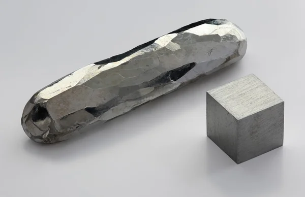 Photographie d'un spécimen de cadmium présentant un cristal métallique facetté allongé à côté d'un petit cube métallique lisse sur une surface grise claire.