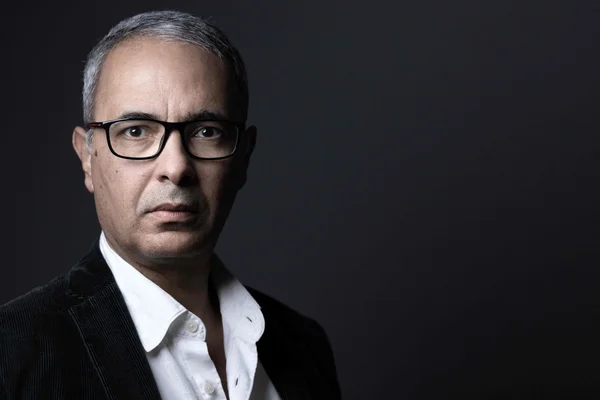 Portrait studio de Kamel Daoud, lauréat du Goncourt 2024, homme d'âge mûr aux cheveux grisonnants et lunettes à monture noire, portant chemise blanche ouverte et blazer de velours côtelé sombre sur fond sombre.