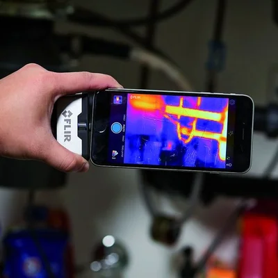 Main tenant un smartphone équipé d'une caméra thermique Flir One de Teledyne Flir, affichant une carte de chaleur de tuyaux en arrière-plan.