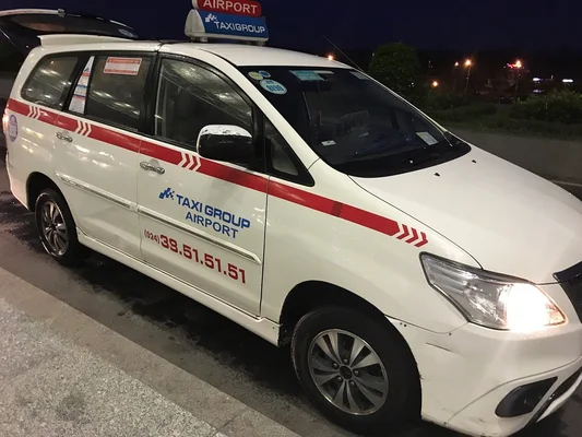 Taxi de l'aéroport de Hanoï, Toyota Avanza blanc à bandes rouges et marque 'TAXI GROUP AIRPORT', stationné de nuit avec ses phares allumés.
