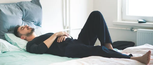 Méditer dans son lit et pleine conscience MBSR : un homme barbu est allongé sur le dos sur un lit, yeux fermés, mains croisées sur le ventre, en tenue sombre près d'une fenêtre lumineuse.