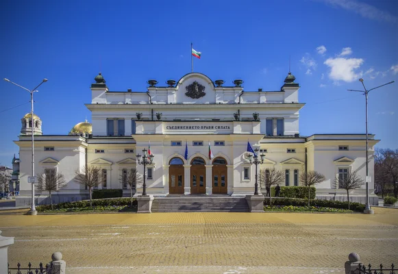 Façade du bâtiment de l'Assemblée nationale de Bulgarie à Sofia, avec fronton central orné d'un blason, drapeau bulgare et l'inscription 'СЪЕДИНЕНИЕТО ПРАВИ СИЛАТА', symbole du parlement en jeu lors des législatives.