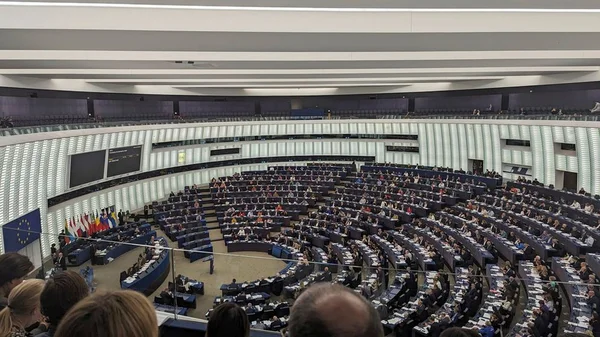 Le Parlement européen, vu de haut, avec des rangées de bureaux courbes occupés par des députés et un drapeau européen à gauche, dénonçant l'incitation à la haine dans les manuels scolaires palestiniens et de l'UNRWA.