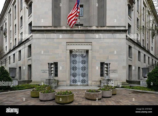 Façade du bâtiment du département de la Justice Robert F. Kennedy à Washington D.C., avec un grand drapeau américain au-dessus d'une porte en métal décorative et des jardinières en pierre à l'avant-plan.