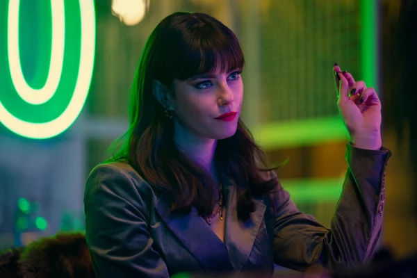 Personnage féminin de la série The Gentlemen (2024) de Guy Ritchie : femme aux cheveux foncés et frange, en blazer gris, assise dans une pièce sombre éclairée par des néons verts, tenant un petit objet levé.