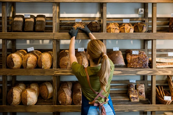Dans le cadre de l'autorisation des boulangeries et fleuristes artisanaux à faire travailler leurs salariés ce 1er mai, une femme blonde en queue-de-cheval, t-shirt vert et tablier bleu, range des pains sur une étagère en bois en boulangerie.