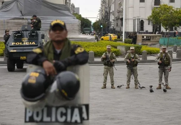 Soldats et policiers montant la garde à la Plaza San Martín à Lima, au Pérou, le 16 décembre : policier flou en premier plan avec un bouclier, trois soldats en camouflage à l'arrière près d'un taxi jaune et d'un véhicule blindé.