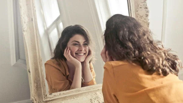 Photographie de l'article « Pourquoi les visages paraissent différents dans un miroir et une photo ? » de Caminteresse.fr : une femme aux cheveux bouclés, en haut mustard, souriant à son reflet dans un miroir à cadre ornementé et usé, menton sur les mains.