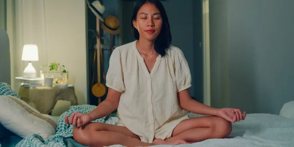 Beditation selon Marie Claire : une femme aux cheveux foncés médite assise en tailleur sur son lit, yeux fermés, mains sur les genoux, dans une chambre tamisée avec lampe et chapeaux.