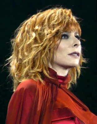 Mylène Farmer de profil, aux cheveux bouclés roux-blonds et portant un vêtement rouge à col haut, sur fond sombre, regardant vers le haut.