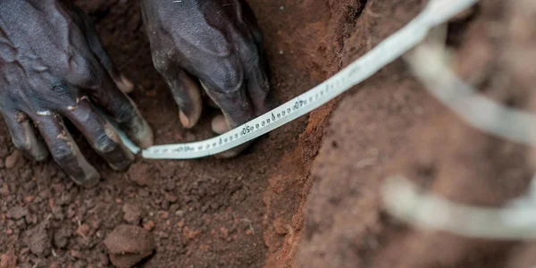 Gros plan sur des mains utilisant un mètre ruban pour mesurer la profondeur d'un trou dans un sol brun rougeâtre, une démarche de l'Alliance Bioversity International - CIAT sur la santé des sols.