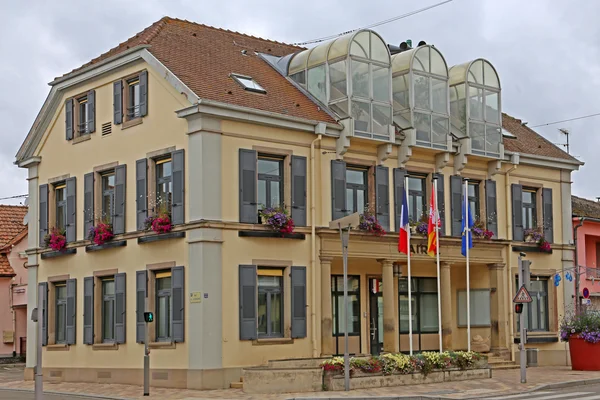 Hôtel de ville de Fessenheim (Haut-Rhin, France), bâtiment jaune à deux étages à toit de tuiles brunes et structure vitrée, drapeaux devant l'entrée, commune connue pour sa centrale nucléaire controversée.