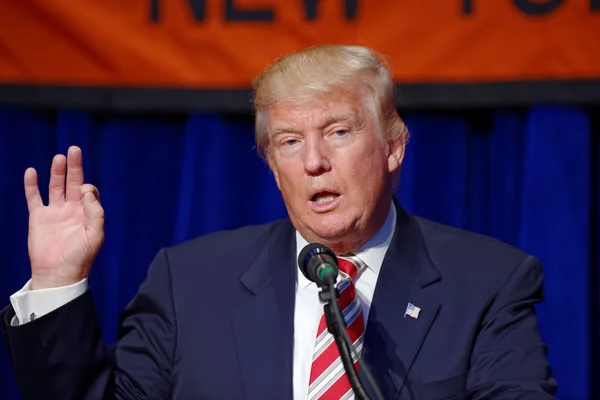 Donald John Trump, homme d'affaires et politicien américain, s'exprimant au pupitre lors d'un événement, portant un costume sombre et une cravate à rayures rouges, la main droite levée.