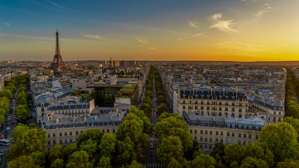 Vue de Paris depuis l'Arc de Triomphe, photographie en plongée avec la tour Eiffel à gauche et de nombreux immeubles haussmanniens sous un ciel de coucher du soleil, le tout encadré par des barres grises.
