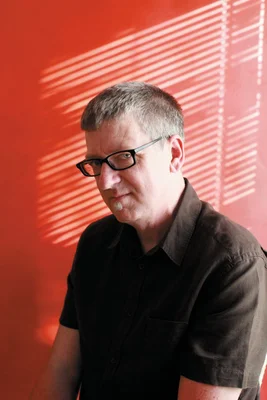 Backderf, auteur de bandes dessinées, homme d'âge mûr aux cheveux gris courts et bouc, portant lunettes à monture noire et chemise noire à manches courtes, regardant hors champ sur fond rouge à motifs d'ombre diagonaux.