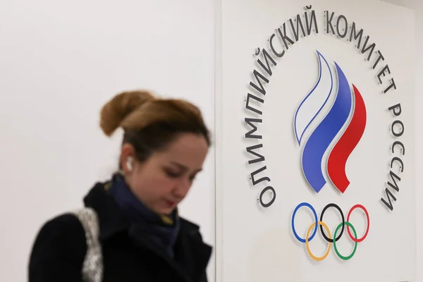 Femme au chignon marchant devant un mur affichant le logo du Comité olympique russe, une flamme stylisée aux couleurs de la Russie sur les anneaux olympiques, alors que se pose la question de la présence russe aux Jeux de Milan 2026.