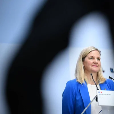 Femme blonde en blazer bleu parlant à un podium muni de micros devant un fond abstrait flou, lors d'une intervention sur la décision du CIO de réintégrer les athlètes russes avant les JO de Los Angeles.