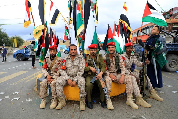 Cinq hommes en uniformes militaires, identifiés comme rebelles houthis du Yémen promettant de continuer leurs attaques contre Israël, sont assis sur une barrière en béton jaune devant de grands drapeaux palestiniens, avec un homme en tenue traditionnelle tenant un drapeau.
